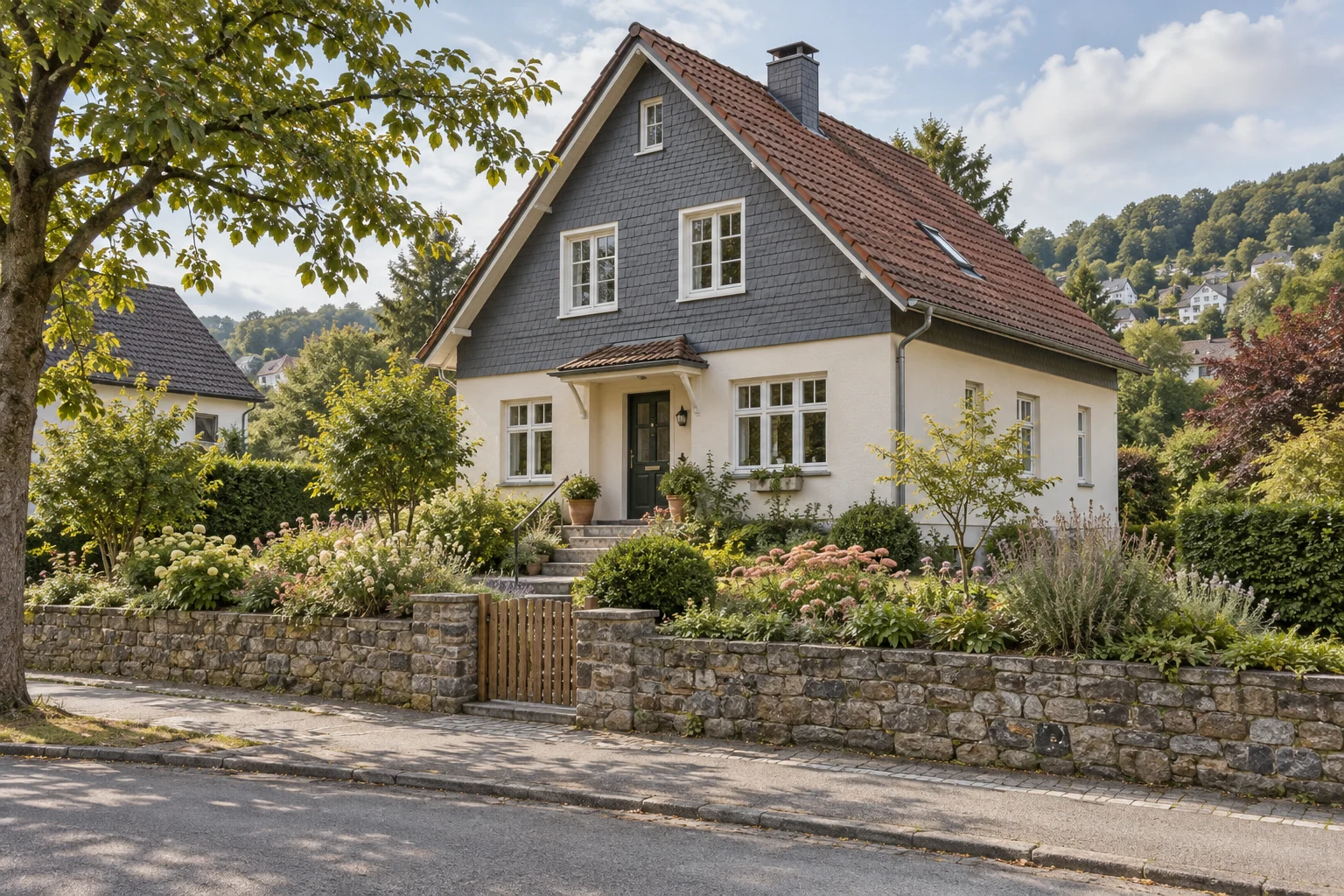 Helles Einfamilienhaus mit dunklem Schiefergiebel und braunrotem Ziegeldach hinter einer Natursteinmauer mit Gartentor in grüner Hanglage.