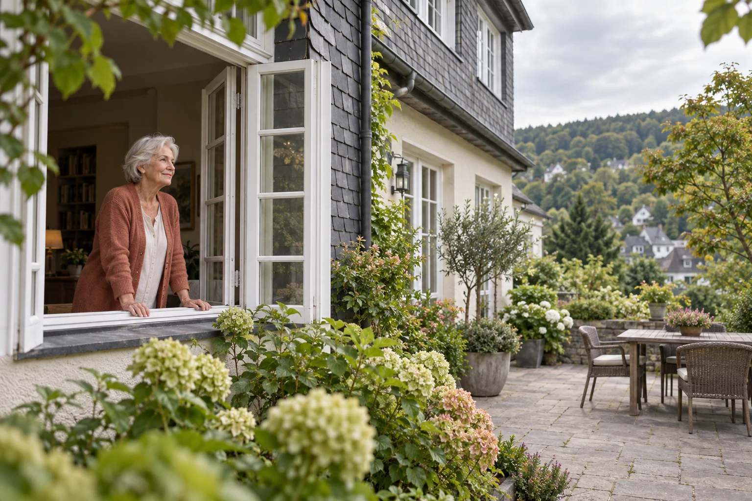 Ältere Frau mit silbernem Haar und terrakottafarbener Strickjacke steht am offenen Fenster eines Hauses mit Schieferfassade und blickt in den grünen Garten.